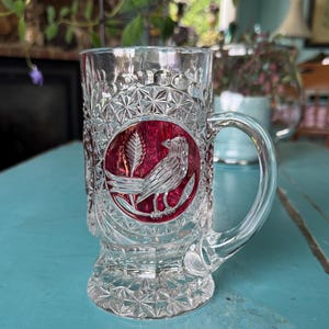 May include: Clear glass beer mug with a handle and a decorative etched pattern. The mug features a red circular emblem with a detailed bird and wheat design. The glass has a faceted base and a clear handle.