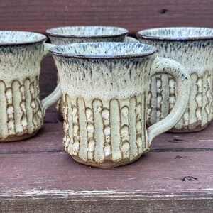 May include: A set of three ceramic mugs featuring a cream-colored glaze with a dark blue drip pattern around the rim. Each mug has a textured exterior with vertical indentations and a sturdy handle, showcasing a handcrafted appearance.