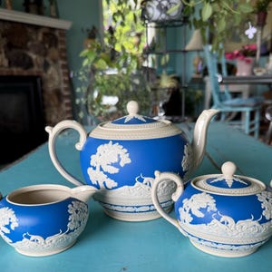 May include: A blue and white ceramic tea set, including a teapot, sugar bowl, and creamer. The set features a raised white relief design of trees and figures against a blue background. The teapot has a curved spout and handle.