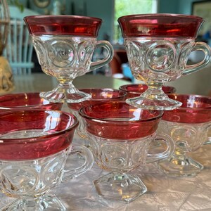 May include: A collection of vintage clear glass teacups and saucers with a ruby red rim. The teacups have a decorative pattern and a handle. The saucers are round. The teacups and saucers are arranged on a white tablecloth.