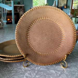 May include: A set of four brown ceramic plates with a textured surface and a gold-colored rim. The plates are displayed on a gold-colored metal stand. The plates have a decorative dotted pattern around the edge.