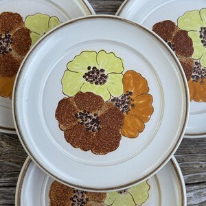 May include: A set of four round ceramic plates featuring a cream-colored base with a brown rim. Each plate displays a central floral motif consisting of three stylized flowers in shades of light green, dark brown, and golden orange with dark centers.