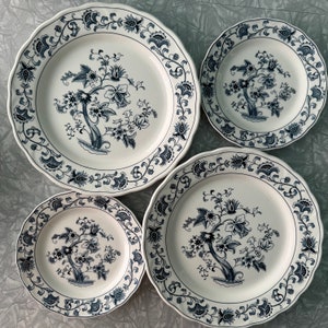 May include: Set of four white ceramic dinner plates with a blue floral design. The plates have a scalloped edge and a delicate floral pattern with a tree in the center.