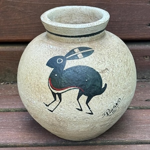 May include: A round, textured beige ceramic vase featuring a hand-painted black rabbit silhouette with a red and white stripe detail. The vase has a wide opening with a thin black line around the neck and the signature Tuland NEI written in black.
