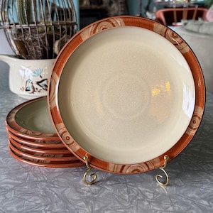 May include: A set of six cream-colored ceramic plates with a rust-colored border featuring a wood-grain design. One plate is displayed on a gold-colored stand. The plates have a simple, elegant design, suitable for everyday use or special occasions.