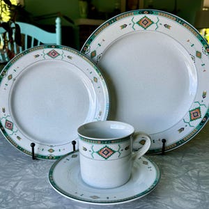May include: A set of three white ceramic plates with a green, brown, and orange geometric pattern. The plates are in different sizes and are arranged in a pyramid shape. The smallest plate is a saucer for a cup.