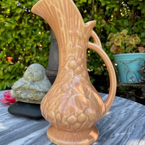 May include: A peach-colored ceramic pitcher with a floral design and a handle. The pitcher has a wide mouth and a rounded base.