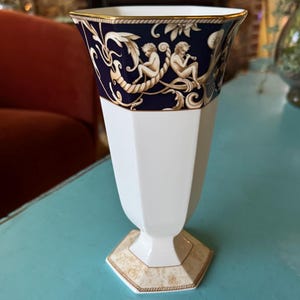 May include: White and navy blue decorative vase with a gold rim. The vase features a detailed design of cherubs and foliage. The base has a gold and beige geometric pattern. The vase is on a light blue surface.
