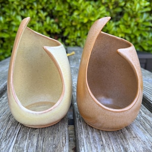 May include: Two teardrop-shaped ceramic yarn bowls with a large side opening for yarn. One bowl is a pale cream color, and the other is a warm terracotta orange. Both feature a smooth, matte finish and a curved, asymmetrical rim design.