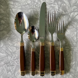 May include: A set of five stainless steel and brown handled silverware pieces. The set includes a large spoon, a small spoon, a knife, and two forks. The handles are a dark brown color with gold-colored accents at the base and top.