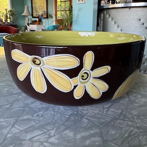 May include: A large, decorative ceramic bowl with a glossy brown exterior and a light green interior. The bowl features a floral design with yellow and white daisy-like flowers. The bowl is suitable for serving or display.