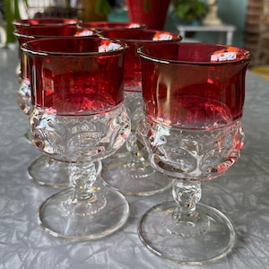 May include: Set of six clear glass stemmed drinking glasses with a ruby red rim and a clear, textured base. The glasses are arranged in a row on a white surface.