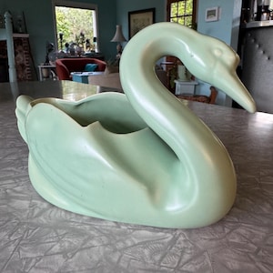 May include: A pale green ceramic swan planter. The swan has a long, curved neck and a detailed beak. The planter has a large opening for plants or other decorative items. The swan is a classic decorative piece.
