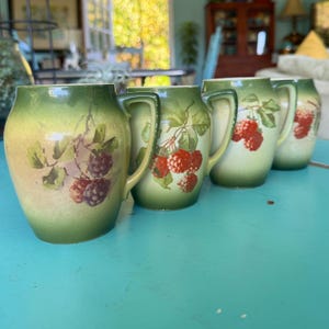May include: Four antique ceramic pitchers with a green and yellow ombre glaze. Each pitcher features a hand-painted design of red raspberries and green leaves. The pitchers have a curved handle and a rounded body.