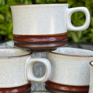 May include: A stack of ceramic mugs featuring a speckled off-white upper section and a solid dark brown glazed base. Each mug has a wide, rounded handle and a distinct horizontal ridge separating the two-toned colors on the exterior surface.