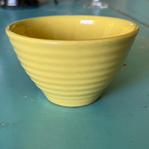 May include: A yellow ceramic bowl with a ribbed exterior. The bowl has a rounded shape and is sitting on a turquoise surface. The bowl is empty and appears to be clean.
