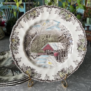 May include: A stack of decorative plates with a winter scene featuring a red covered bridge, snow, and trees. The plates have a white background with a brown floral border and a gold-colored metal stand.
