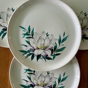 May include: A set of white ceramic plates featuring a hand-painted floral design. Each plate showcases a large white flower with purple accents and green leaves. The plates are arranged on a wooden surface.