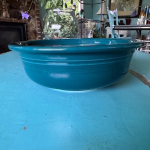 May include: A teal-colored ceramic bowl with a wide rim. The bowl has a smooth, glossy finish and a simple, classic design. The bowl is sitting on a light blue surface.