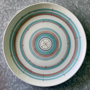 May include: A round ceramic plate featuring a series of concentric circles in shades of turquoise and dusty pink with a crackle glaze finish. The center displays a dark brown starburst motif, and thin dark lines radiate outward across the rings.