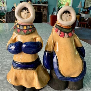 May include: Two ceramic figurines depicting individuals wearing yellow parkas with white fur-trimmed hoods, blue mittens, and dark trousers. Each figure features a colorful geometric collar and a light-colored face with closed eyes and a small mouth.