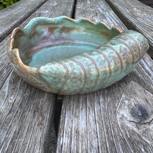 May include: A ceramic bowl shaped like a seashell with a scalloped edge. The interior features a smooth, glossy teal and brown glaze, while the exterior displays a textured, ribbed pattern in shades of green and brown with a matte finish.