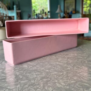 May include: Two rectangular, pale pink containers are stacked on a textured surface. The top container is open, revealing the interior. The containers have a matte finish and are likely made of plastic. The background is blurred.