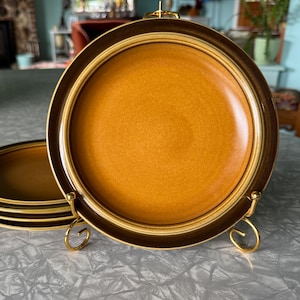May include: A set of vintage ceramic plates with a warm, earthy color palette. The plates feature a textured, speckled surface in shades of brown and gold, with a dark brown border and a gold rim. They are displayed on a gold-colored metal stand.