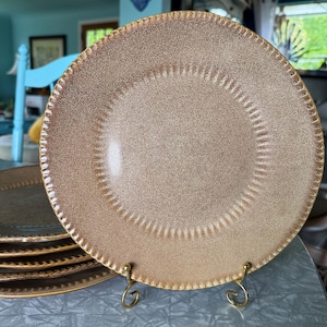 May include: A set of tan-colored ceramic plates with a speckled finish. Each plate has a decorative dotted border and a gold-colored rim. The plates are displayed on a gold-colored metal stand, showcasing their elegant design.
