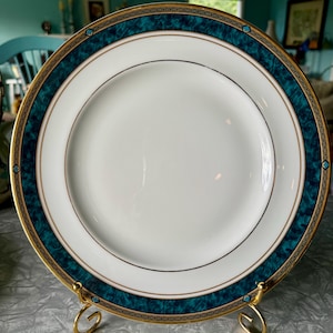 May include: A white ceramic plate with a teal and gold border. The plate has a gold rim and a teal marbled design. The plate is displayed on a gold metal stand. The plate is a piece of dinnerware.