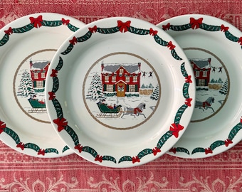 May include: Three white ceramic Christmas plates with a red house, green trees, and a horse-drawn sleigh design. The plates have a green and red garland border with red bows.