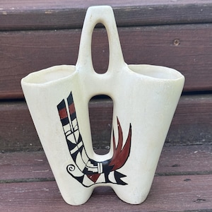 May include: A cream-colored ceramic vase featuring a dual-chamber design with a central handle and open cutout. The exterior is decorated with a hand-painted graphic motif in black, white, and reddish-brown tones, showcasing geometric patterns.