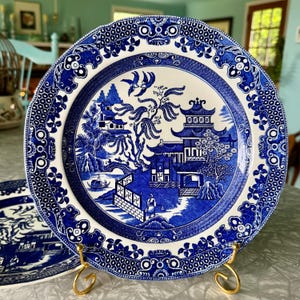 May include: A blue and white ceramic plate with a Willow pattern design. The plate features a detailed landscape scene with pagodas, bridges, and figures. The rim has a decorative border. The plate is displayed on a gold-colored stand.