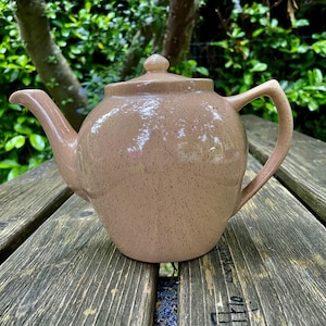May include: A speckled light pink teapot with a rounded body and a curved spout. The teapot has a speckled light pink finish.