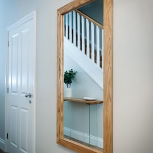 May include: A rectangular mirror with a wooden frame, mounted on a wall. The mirror reflects a staircase with white railings and a small shelf with a vase and a plate. The frame has a natural wood grain.