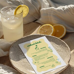 May include: A refreshing drink with ice and a lemon slice sits next to a plate with a weekend details itinerary. The plate is on a sandy surface, with a white cloth and lemon halves in the background. The itinerary lists activities.