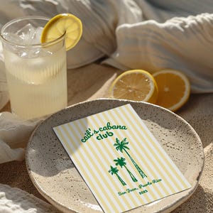May include: A refreshing drink with ice and a lemon slice in a ribbed glass. A plate holds a card with yellow and white stripes, palm tree illustrations, and the text "Cait's Cabana Club, San Juan, Puerto Rico 2023".