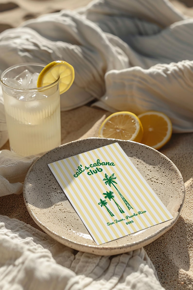 May include: A refreshing drink with ice and a lemon slice in a ribbed glass. A plate holds a card with yellow and white stripes, palm tree illustrations, and the text "Cait's Cabana Club, San Juan, Puerto Rico 2023".