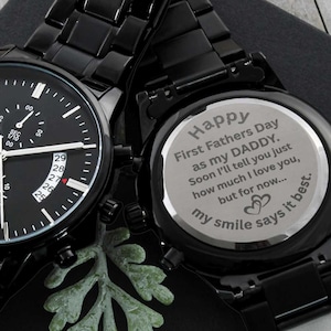 May include: Black wristwatch with a black band and a round face. The watch face has a date window and a small seconds dial. The back of the watch has an engraved message that reads: "Happy First Fathers Day as my DADDY. Soon I'll tell you just how much I love you, but for now... my smile says it best."