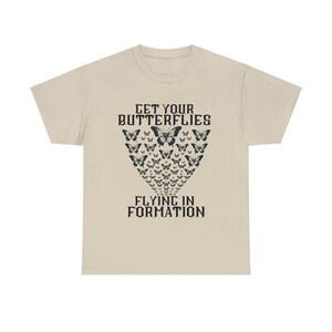 May include: A beige t-shirt with a black graphic design of butterflies flying in formation. The text "Get Your Butterflies Flying In Formation" is printed on the shirt.