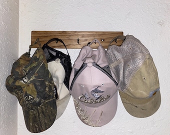 May include: A wooden wall-mounted rack with four metal hooks holding four baseball caps. The caps include a camouflage print, a black and white mesh design, a light pink cap with Cabela's branding, and a tan mesh-back cap with a curved brim.