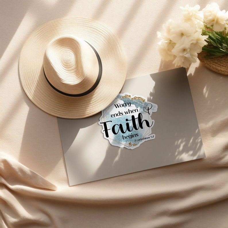 May include: A blue and gold watercolor sticker with the text "Worry ends when Faith begins 2 Corinthians 5:7". The sticker is on a light brown surface with a straw hat and white flowers.