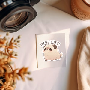 May include: A white card with the text "PUG LIFE" in black lettering. A cartoon pug with brown eyes and a pink nose is sitting on the card.