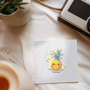 May include: A white card with a yellow pineapple sticker on it. The pineapple has a smiling face and is surrounded by small yellow stars. The card is on a white surface.