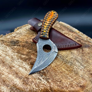 May include: A small, curved knife with a patterned blade and a brown leather sheath. The handle is made of wood and has a yellow and black pattern.