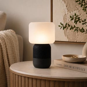 May include: A table lamp featuring a cylindrical black base with a textured mesh middle section and a smooth, rounded, frosted white glass shade that emits a soft, warm glow. The lamp stands on a circular surface next to a book and a small bowl.