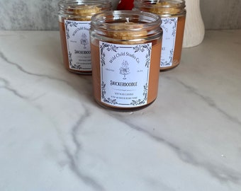 May include: Three glass jars containing brown wax candles. The jars have white labels with the text "Wild Child Studio Co." and "Snickerdoodle" on them. The candles are topped with a brown crumble.