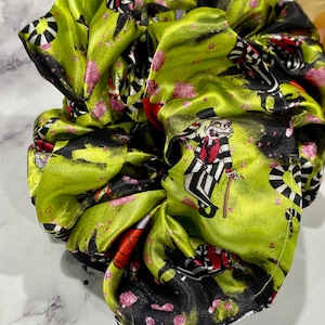 Op de afbeelding: Een groene satijnen scrunchie met een zwart-witte print van een personage uit de film "Beetlejuice".