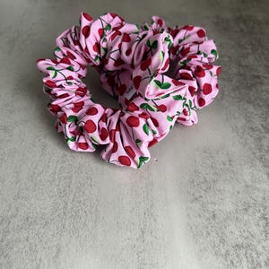 Könnte beinhalten: Zwei rosa Haargummis mit einem sich wiederholenden Muster aus roten Kirschen und grünen Blättern. Diese Haaraccessoires sind aus weichem Stoff gefertigt. Die Haargummis sind so konzipiert, dass sie das Haar sicher halten und einen Hauch von Stil verleihen. Ein niedliches und praktisches Haaraccessoire.