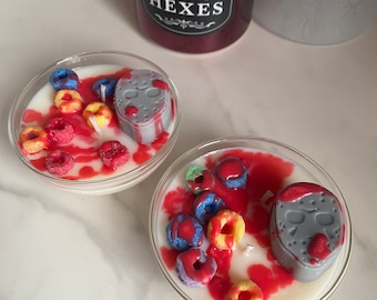 May include: Two glass candle holders with a white wax base, red wax, and colourful cereal pieces. Each holder features a grey wax mask with red accents. The text "hissing HEXES" is visible on a red jar in the background.